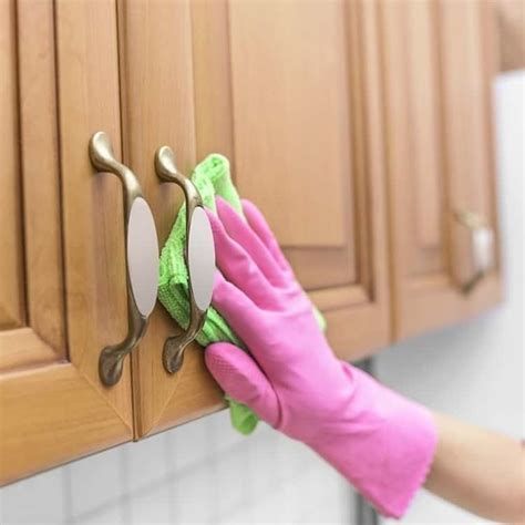 Person wearing pink glove, cleaning a wooden kitchen cabinet with a green cloth.