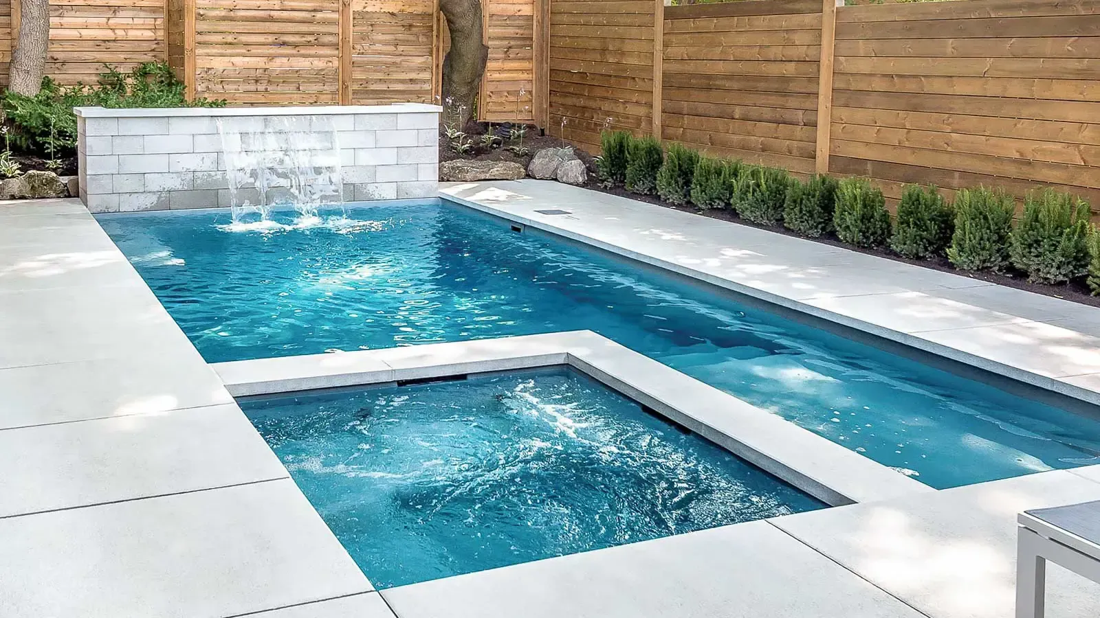 Modern backyard swimming pool with blue water, white patio, wood fence, and small spa area
