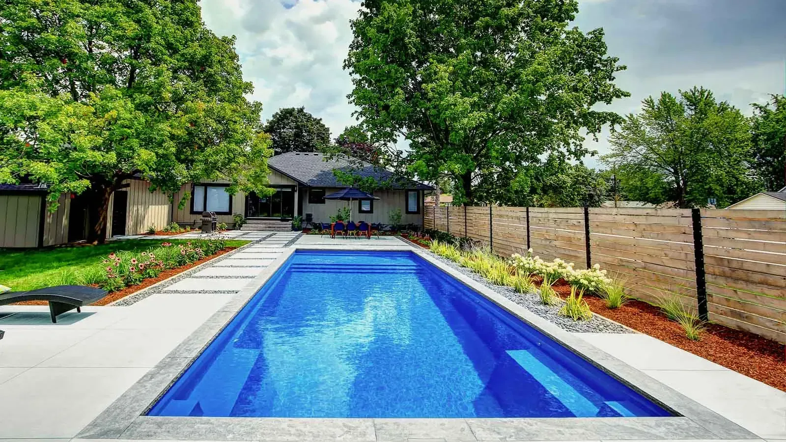 Backyard with a rectangular blue swimming pool, trees, patio, and house in the background