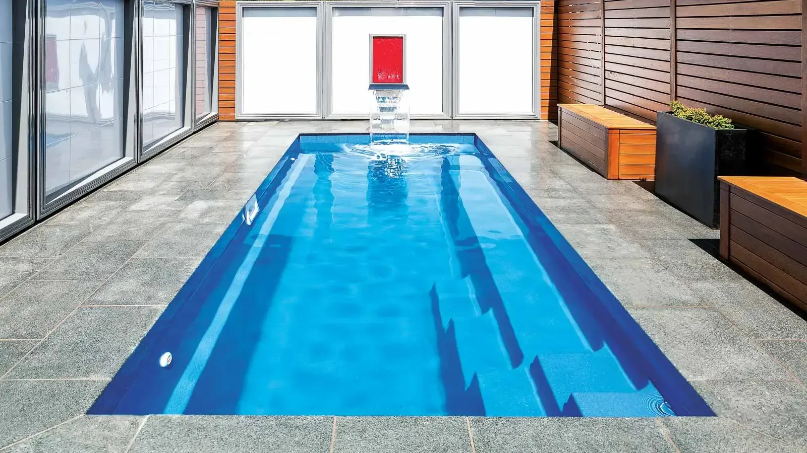 Indoor lap pool with blue water, gray tile deck, and lounge chairs beside a glass wall