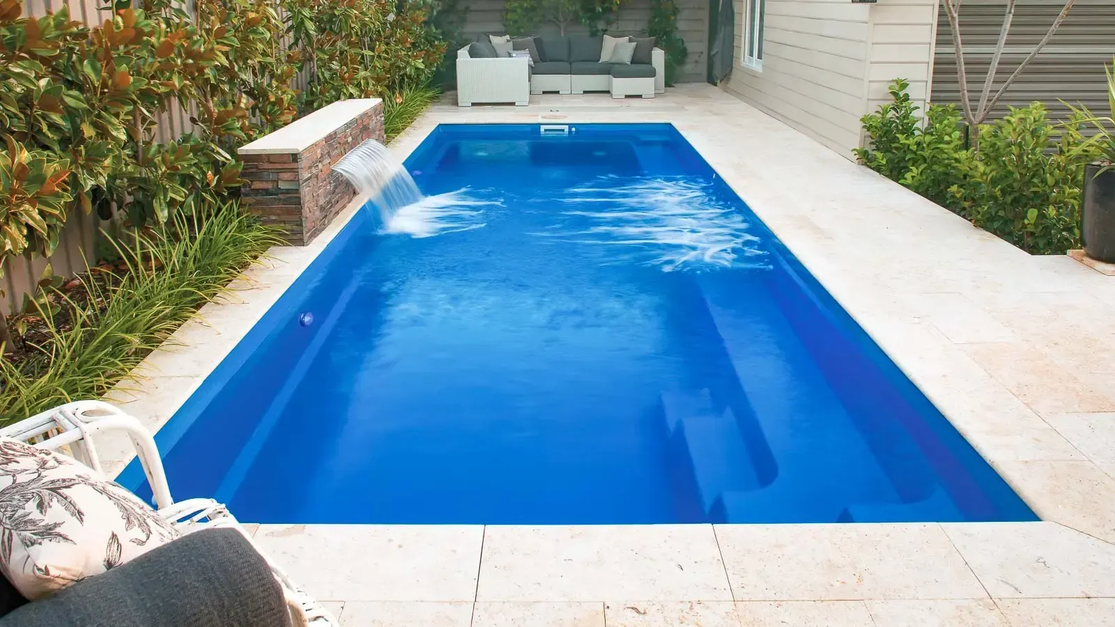 Rectangular backyard swimming pool with bright blue water, surrounded by white stone patio and greenery