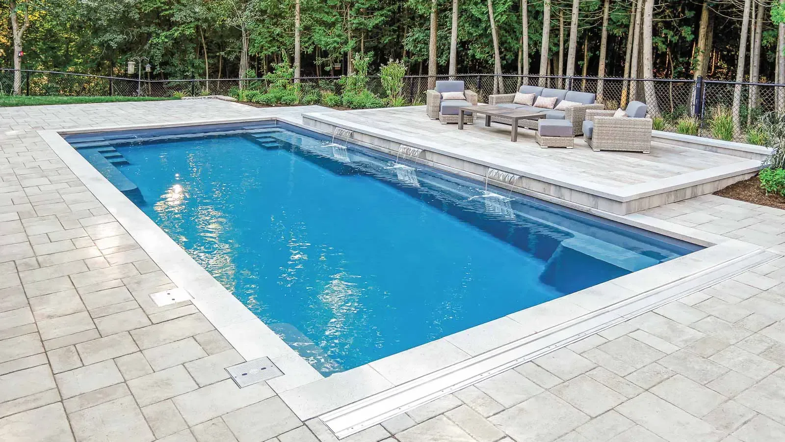 Rectangular blue swimming pool with stone patio and lounge chairs in a wooded backyard