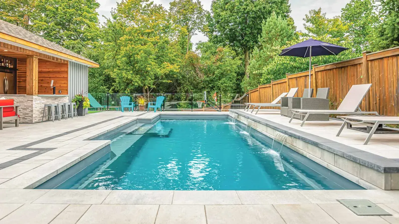 Backyard pool with turquoise water, lounge chairs, umbrella, and a wooden patio surrounded by trees