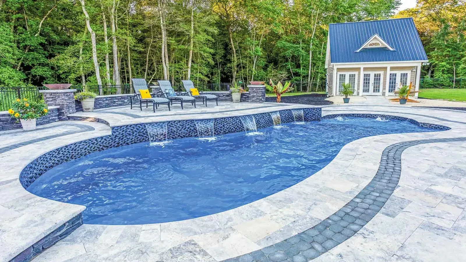 Curved backyard swimming pool with blue water, stone patio, lounge chairs, and a small pool house in the background