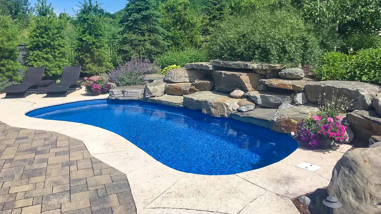 Backyard pool with curved blue water, stone patio, rock waterfall, loungers, and pink flowers amidst greenery