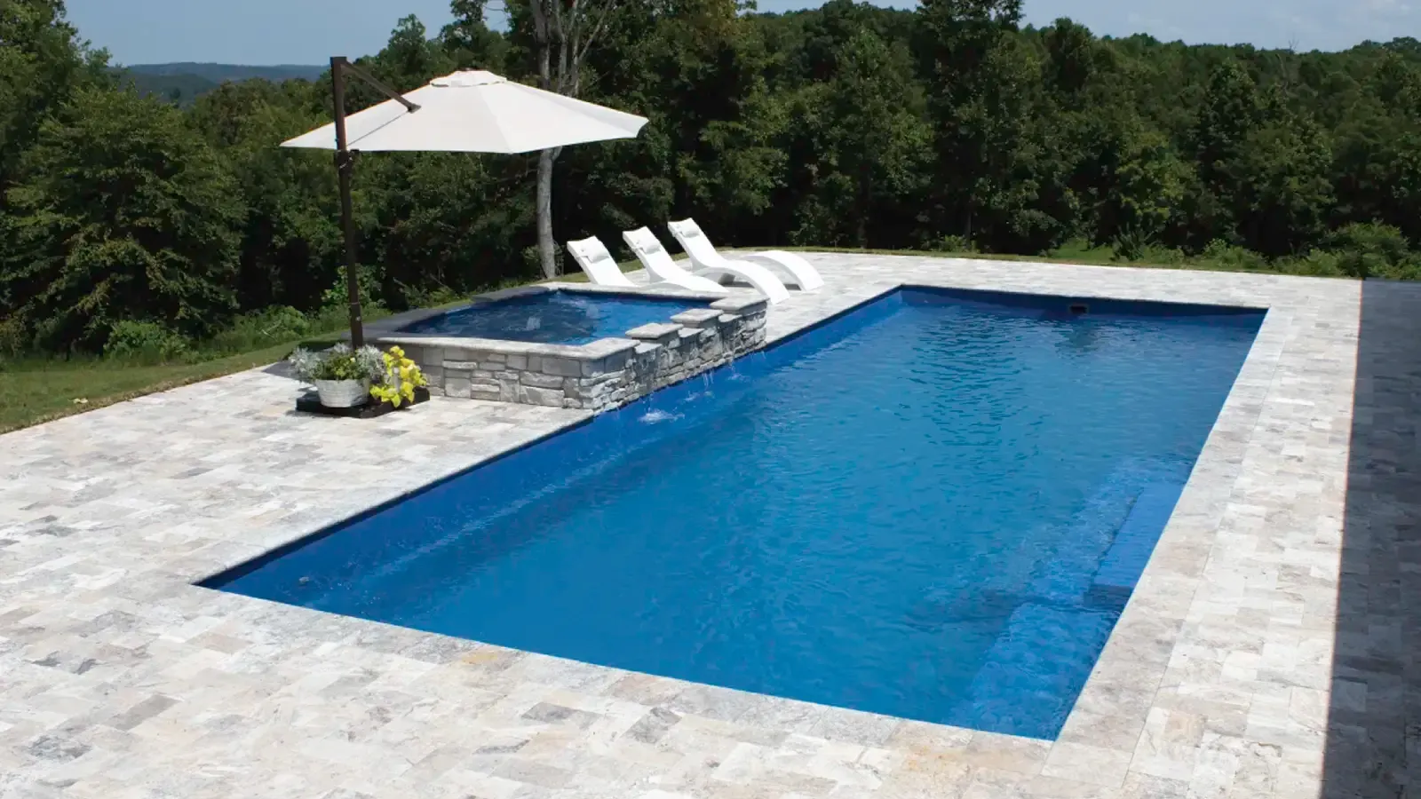 Outdoor pool with blue water, white stone deck, lounge chairs, and a white umbrella overlooking greenery