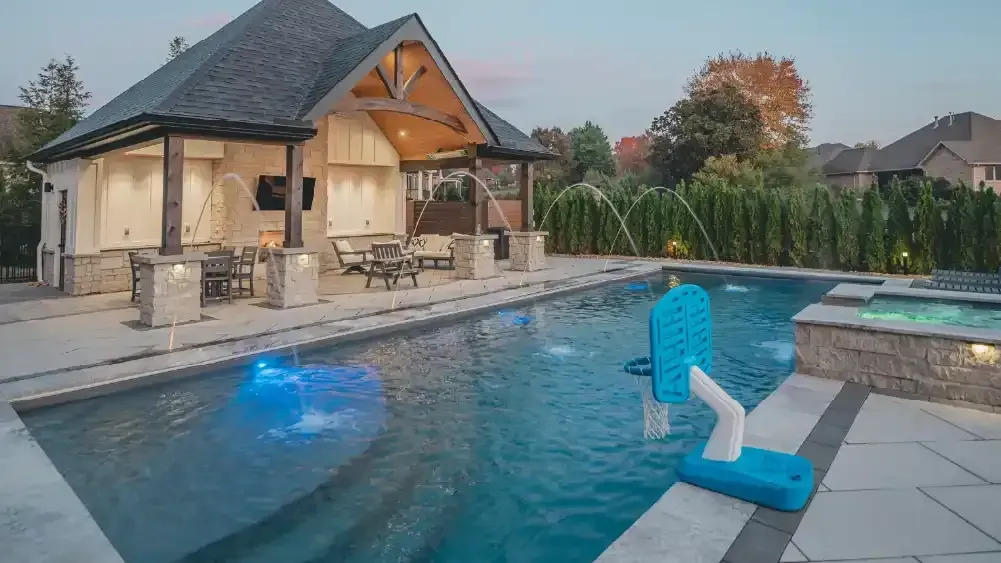 Backyard pool at dusk with a stone patio, lit water, and a blue pool-cleaning robot in the foreground