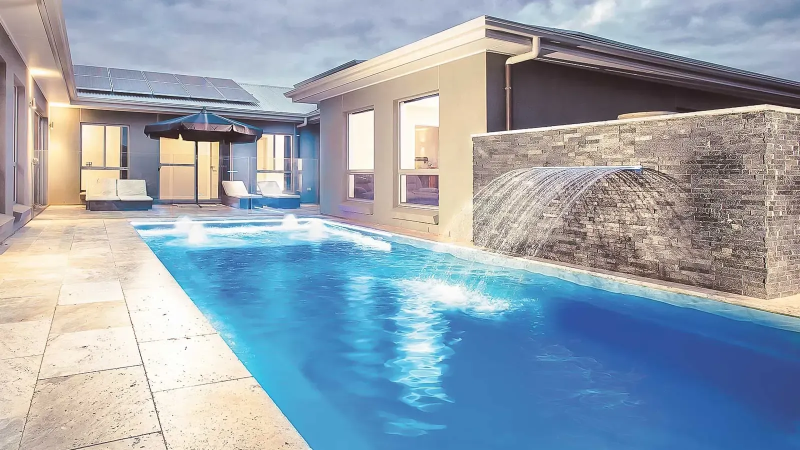 Modern backyard pool with blue water, stone deck, and a small covered patio at dusk
