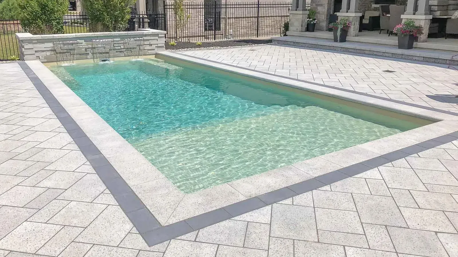 Rectangular outdoor swimming pool with clear turquoise water, surrounded by light stone paving and a metal fence.