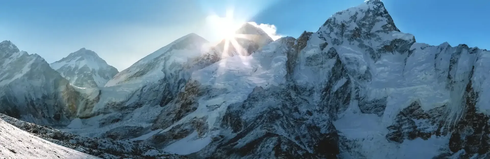 Snowy mountain peaks with the sun rising behind a jagged ridge under a clear blue sky