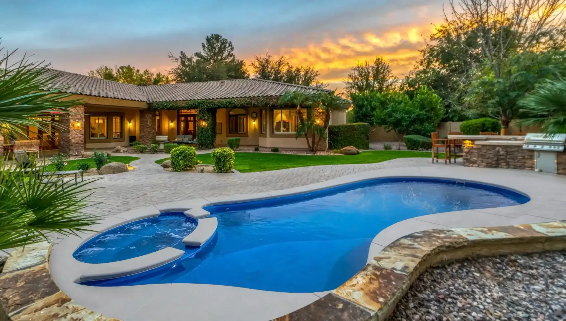 Backyard pool with curved blue water feature at sunset beside a rustic house and patio