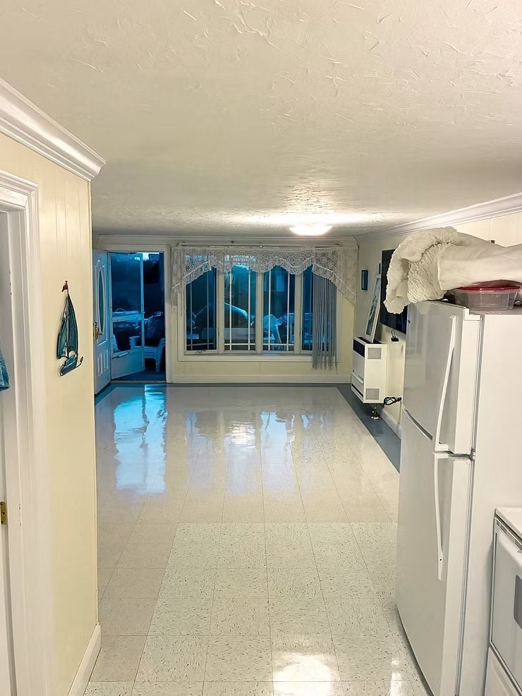 Bright empty kitchen with white cabinets, glossy floor, and large windows at the far end.