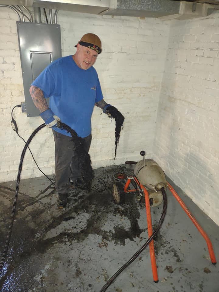 A technician in a blue shirt holds dark sludge pulled from a sewer machine in a white basement.