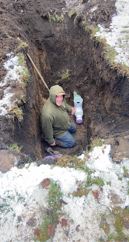 A smiling person in a green hooded jacket kneeling in a muddy, snow-patched trench while installing a PVC pipe fitting.