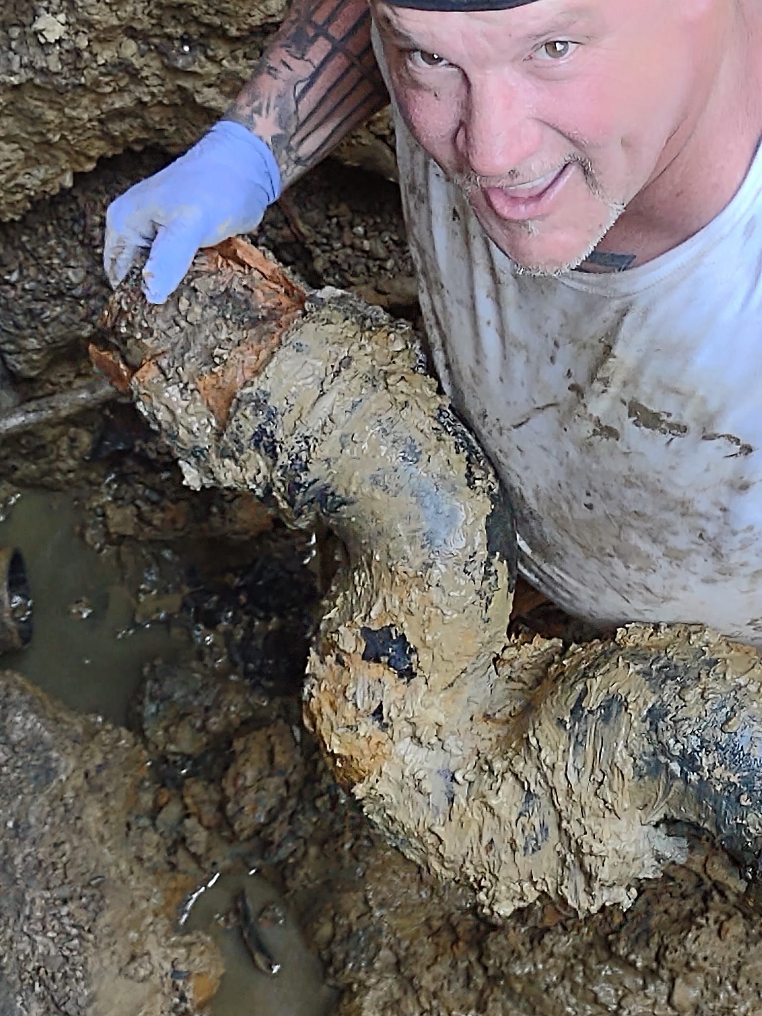 A person wearing a white t-shirt and blue glove smiles while holding a large, muddy pipe section in an excavated trench.