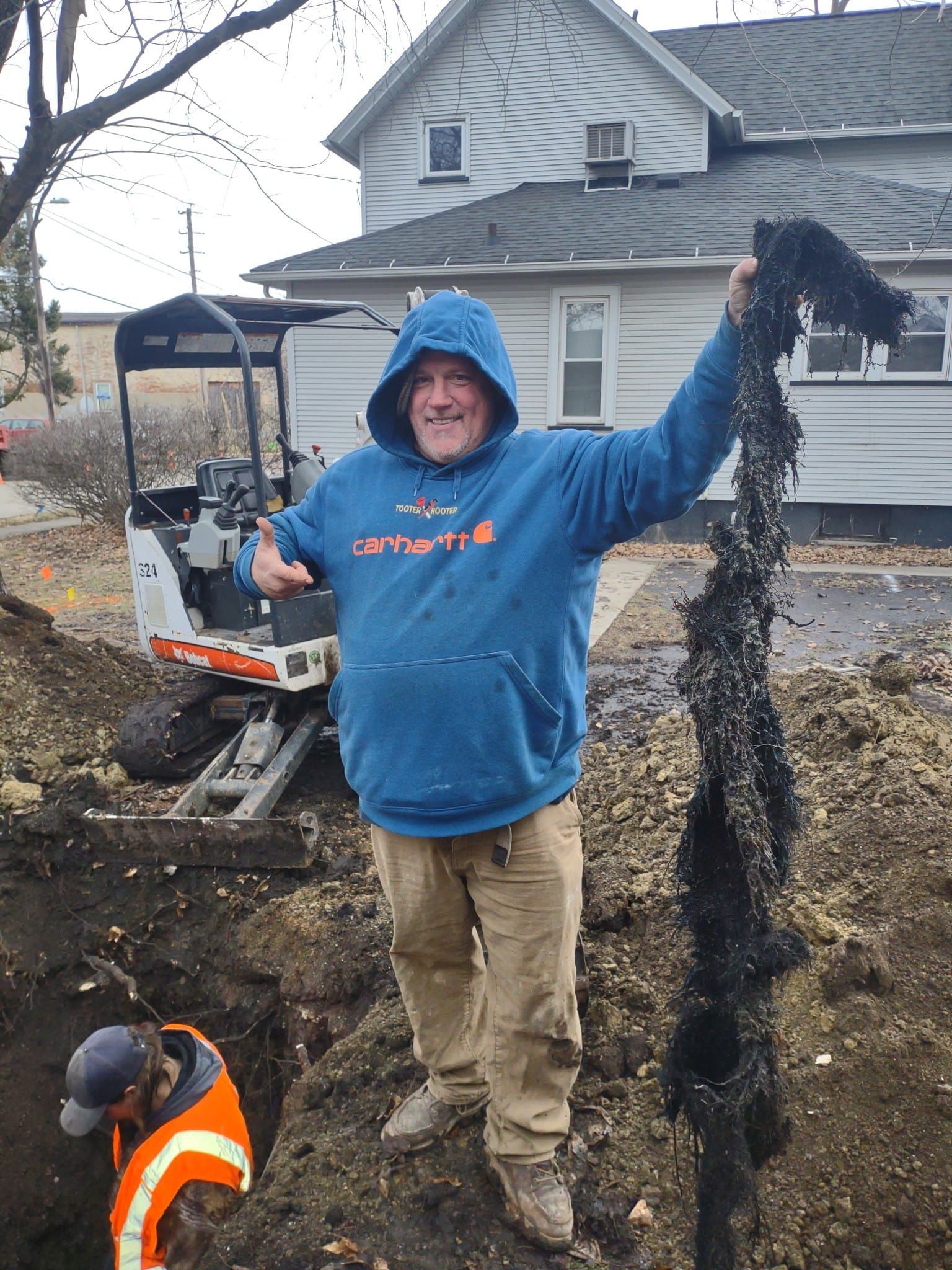 A worker in a blue hoodie stands in a construction site holding a long piece of black debris, with a second worker nearby.