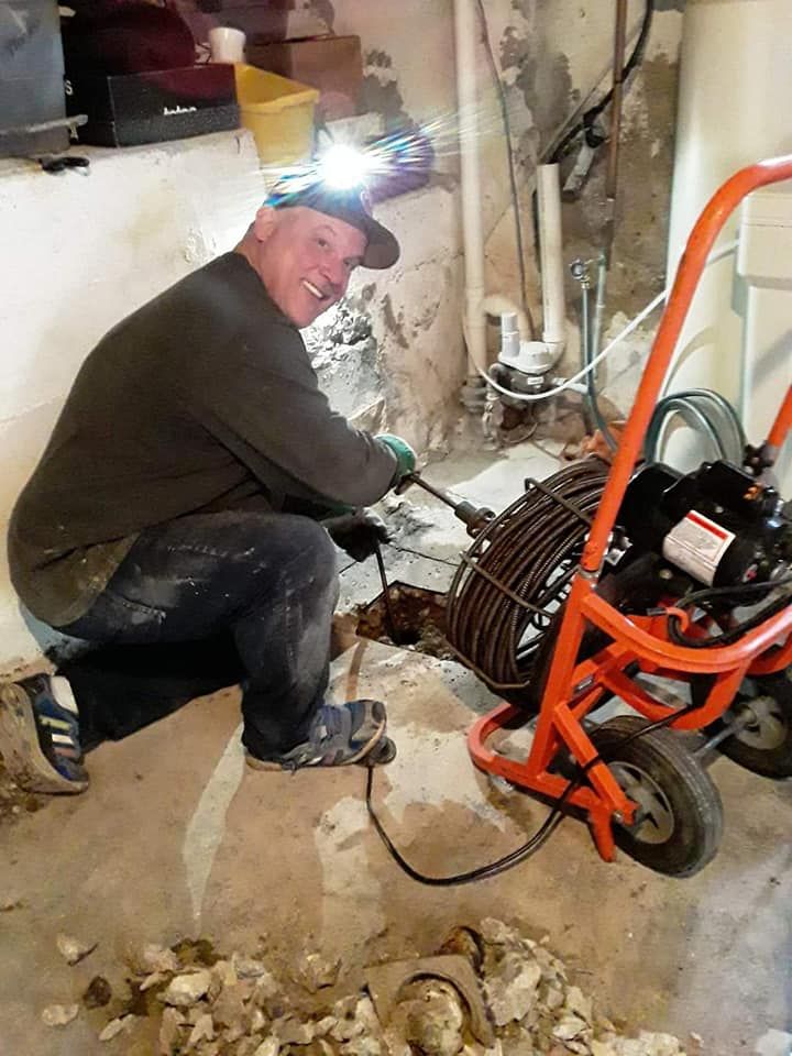 A plumber with a headlamp kneels in a basement, using a mechanical sewer snake to clear a drain in the concrete floor.