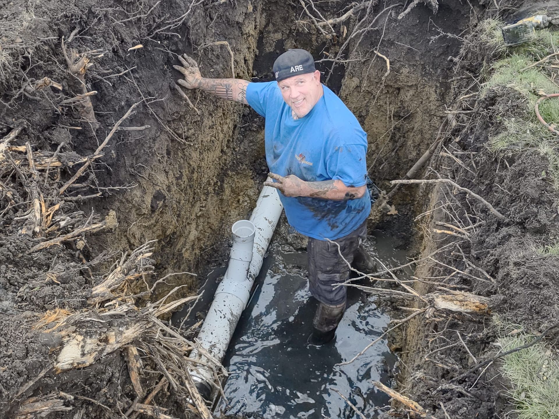 A person in a blue shirt standing in a muddy trench next to a newly installed white sewer pipe.