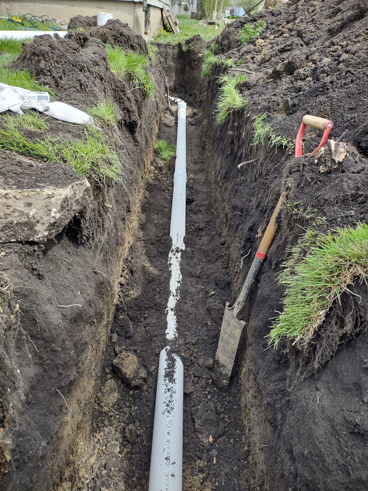 A long, grey PVC pipe runs along the bottom of a narrow, freshly dug dirt trench next to a shovel in a yard.