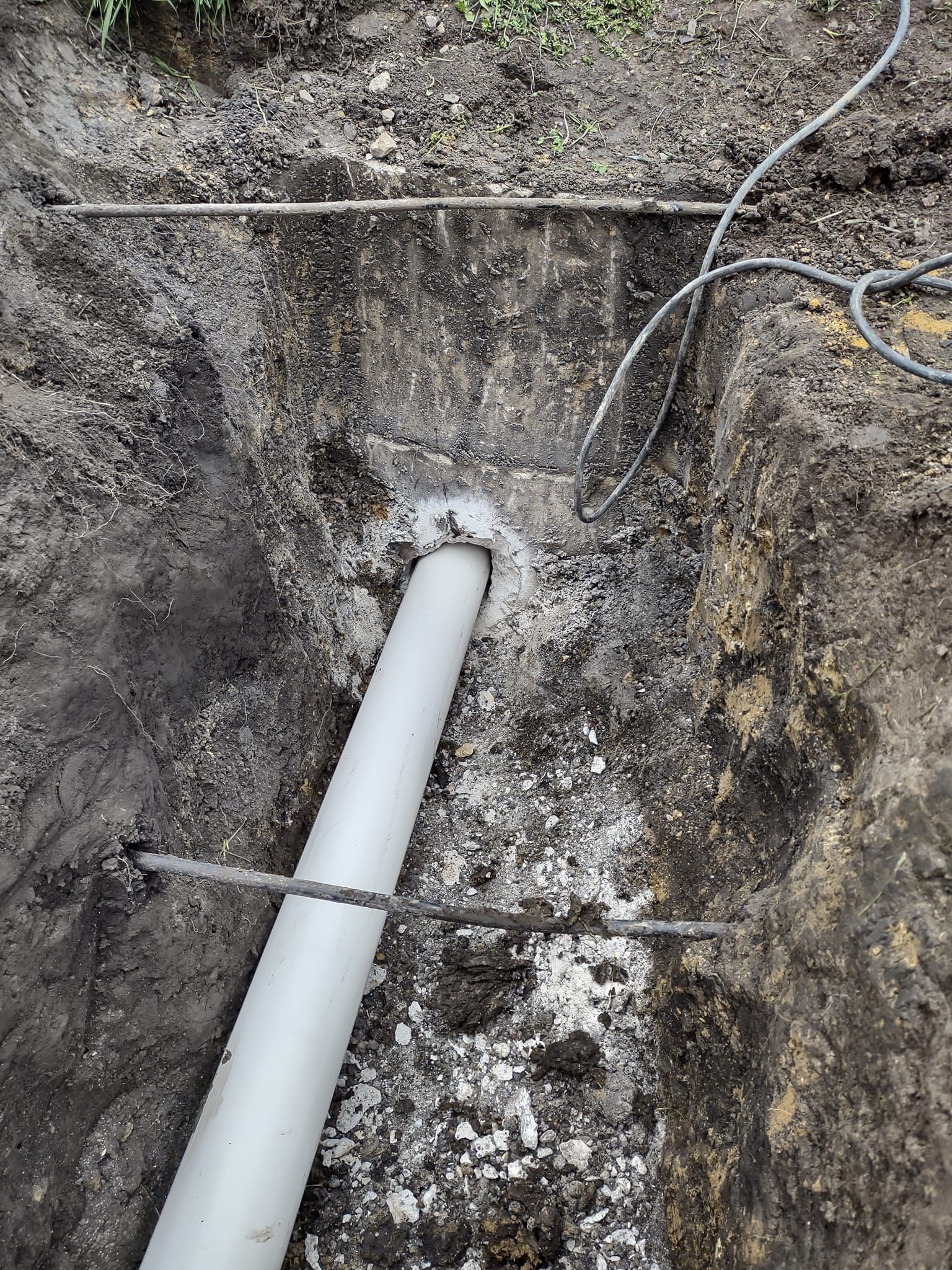 A white PVC pipe extends from a dirt wall into a trench filled with light-colored gravel and reinforcement bars.