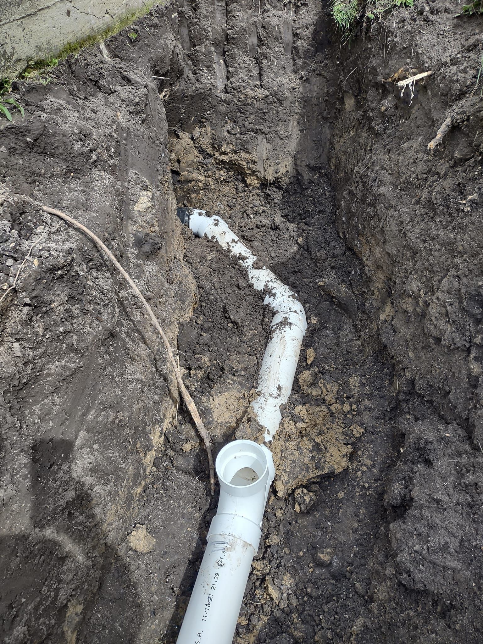 White PVC plumbing pipes joined by an elbow connector in an open dirt trench.