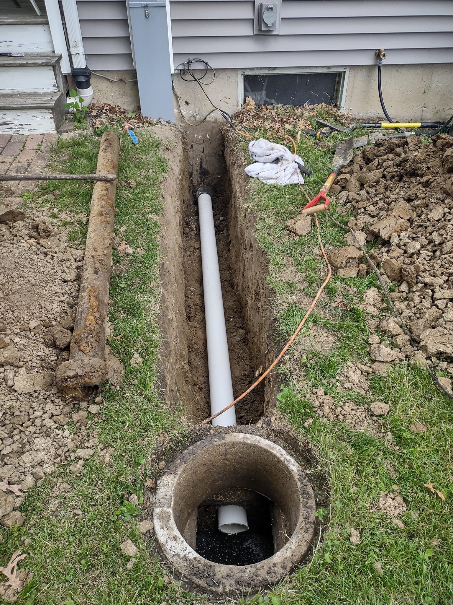 A white PVC pipe runs through an excavated trench in a yard, connecting to a concrete drainage basin in the foreground.