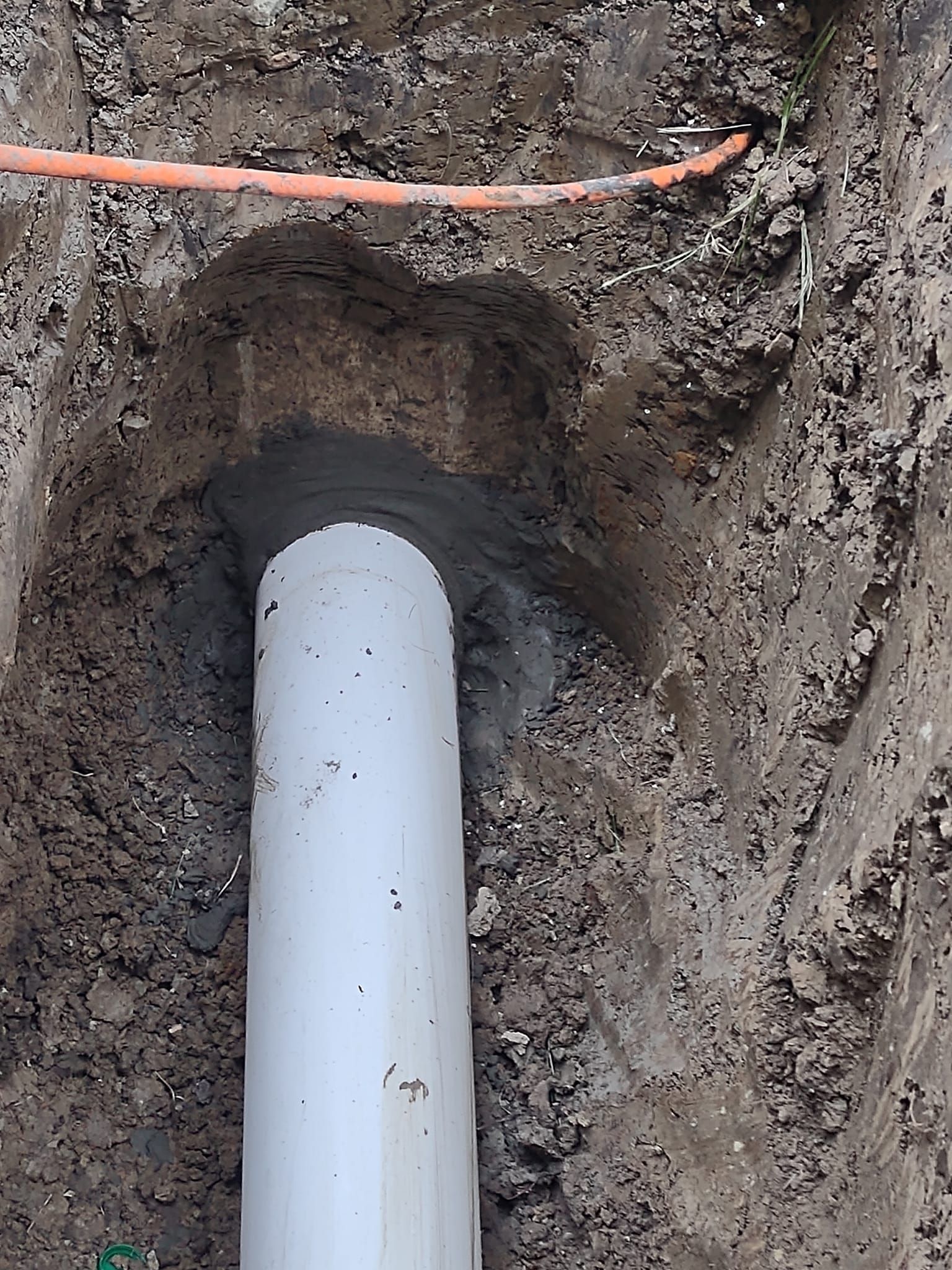 A grey pipe is partially encased in wet concrete at the base of a narrow, excavated dirt trench, below an orange cable.
