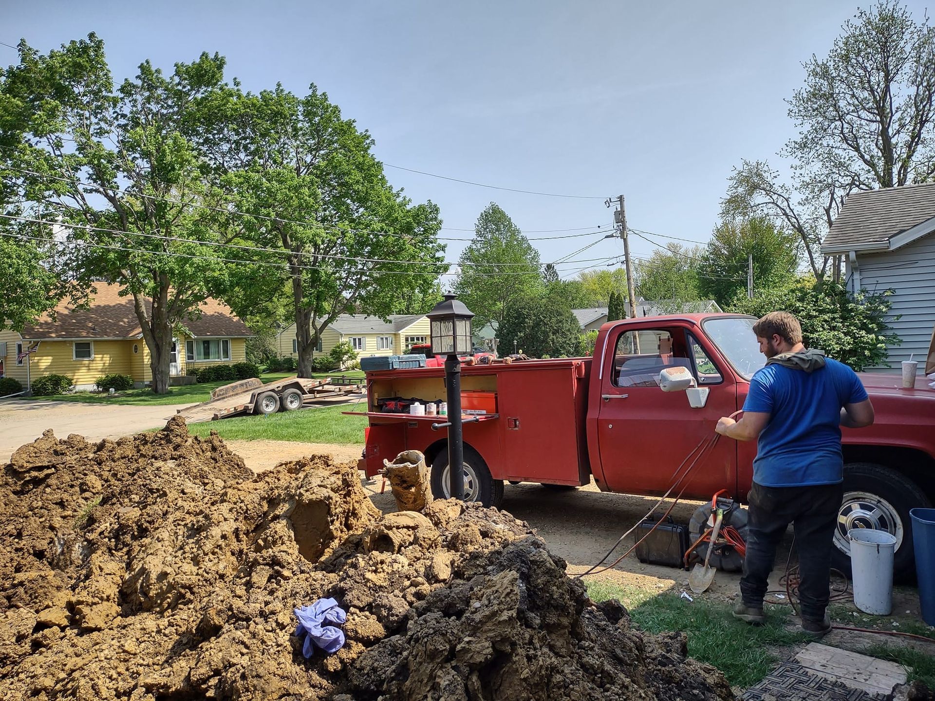 A person in a blue shirt stands by a red utility truck parked near a large pile of dirt in a residential area.