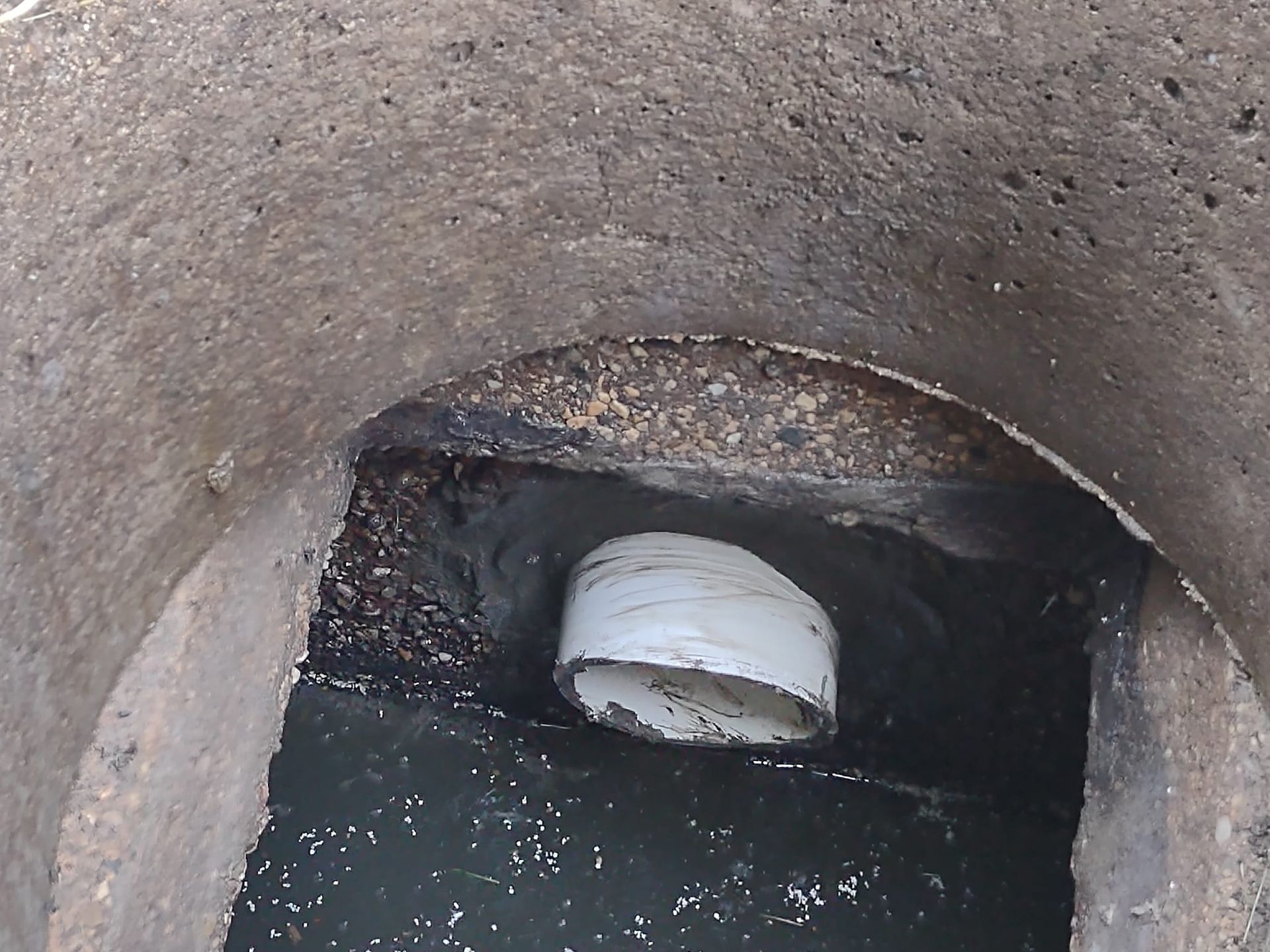 A light-colored plastic pipe protrudes into a dark concrete drainage manhole, sealed with black sealant.