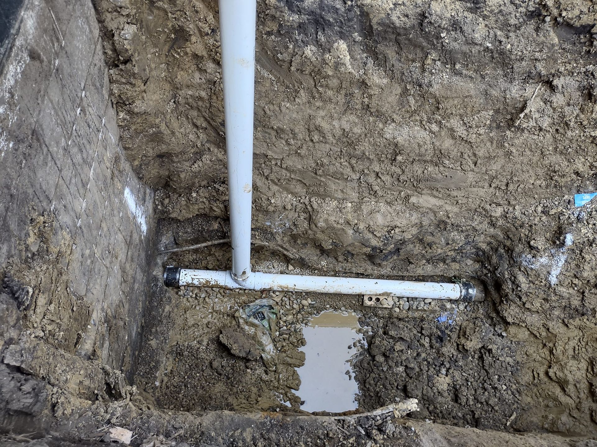 A white PVC T-pipe connection installed in a dirt trench next to a concrete foundation wall.