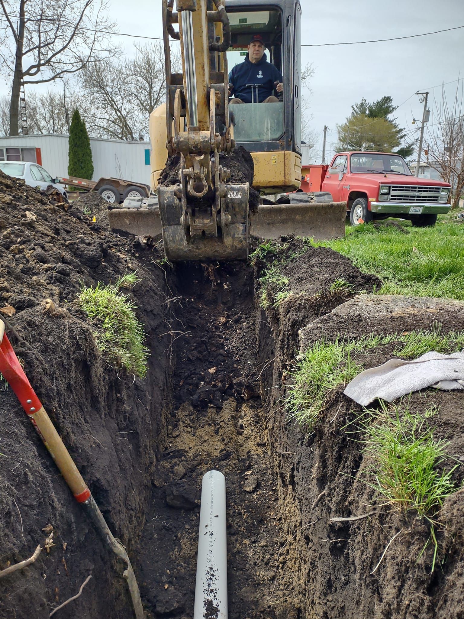 A mini excavator parked over a narrow trench containing a pipe, with a red truck visible in the grassy background.