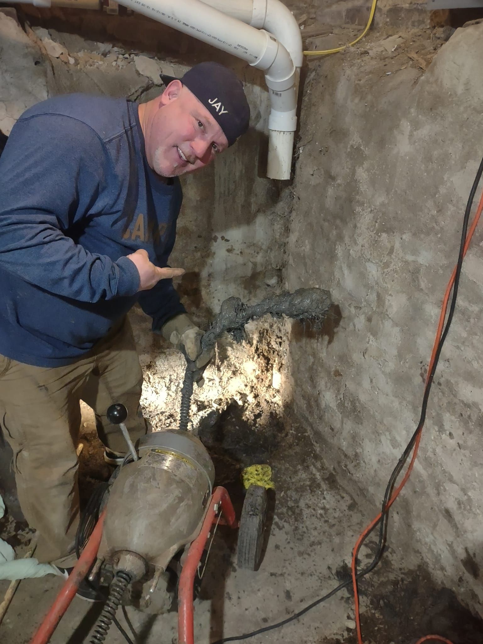 A person using a sewer snake machine to clear a pipe in a basement, pointing toward a messy, sludge-covered cable.