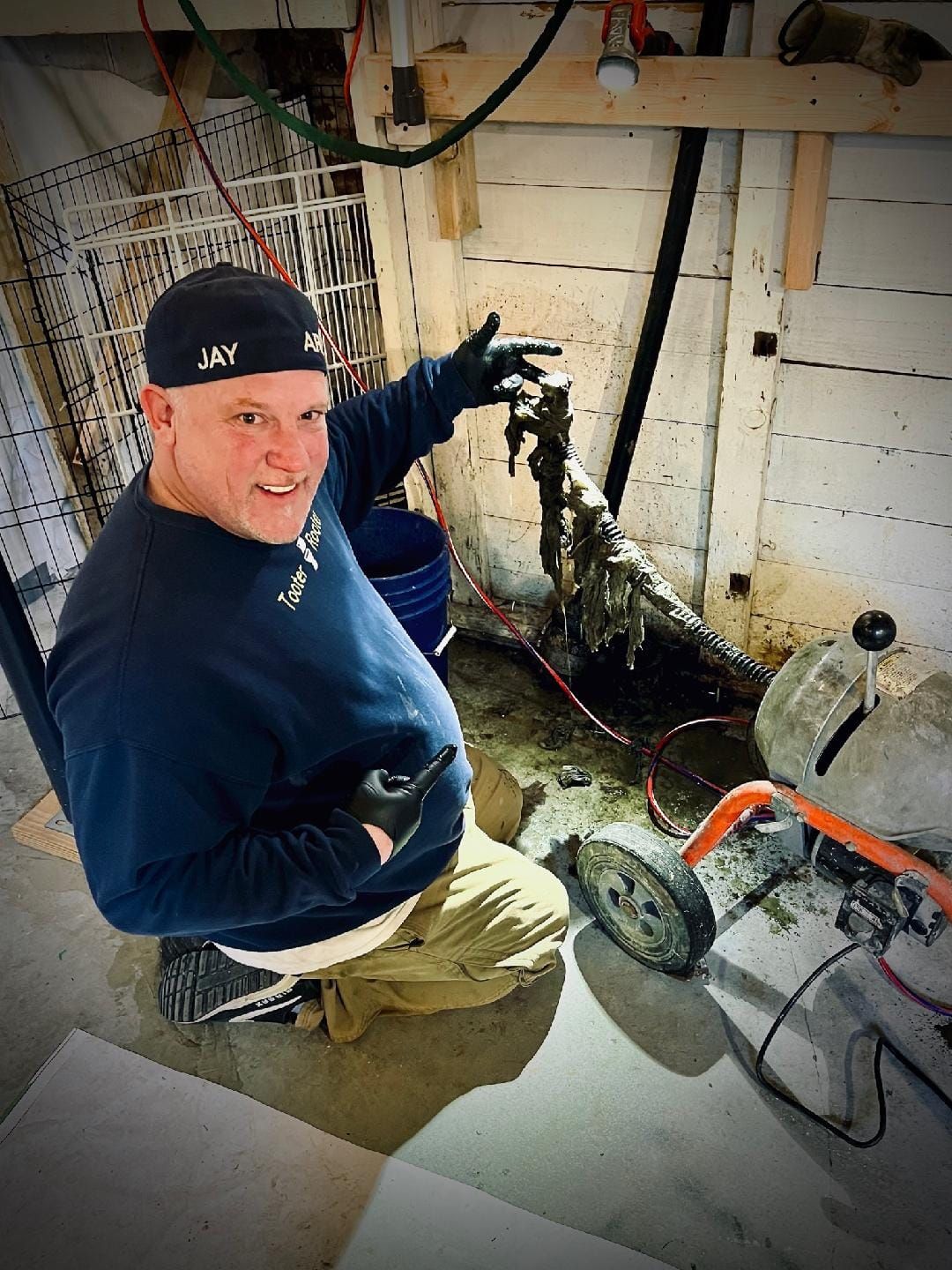 A service technician kneels indoors, pointing toward a piece of plumbing equipment connected to a drain cleaning machine.