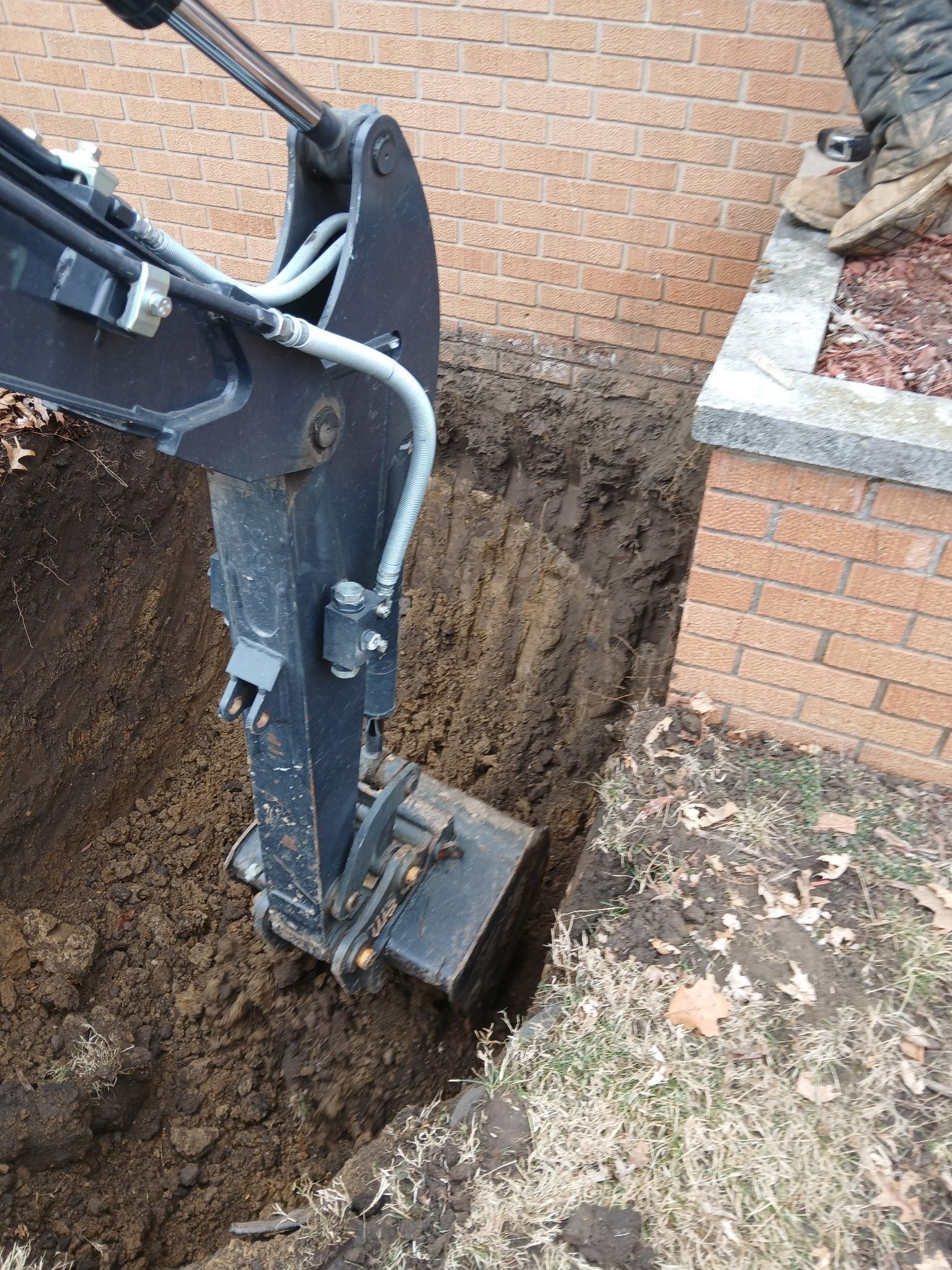 Excavator arm digging a deep trench beside a brick house and retaining wall.