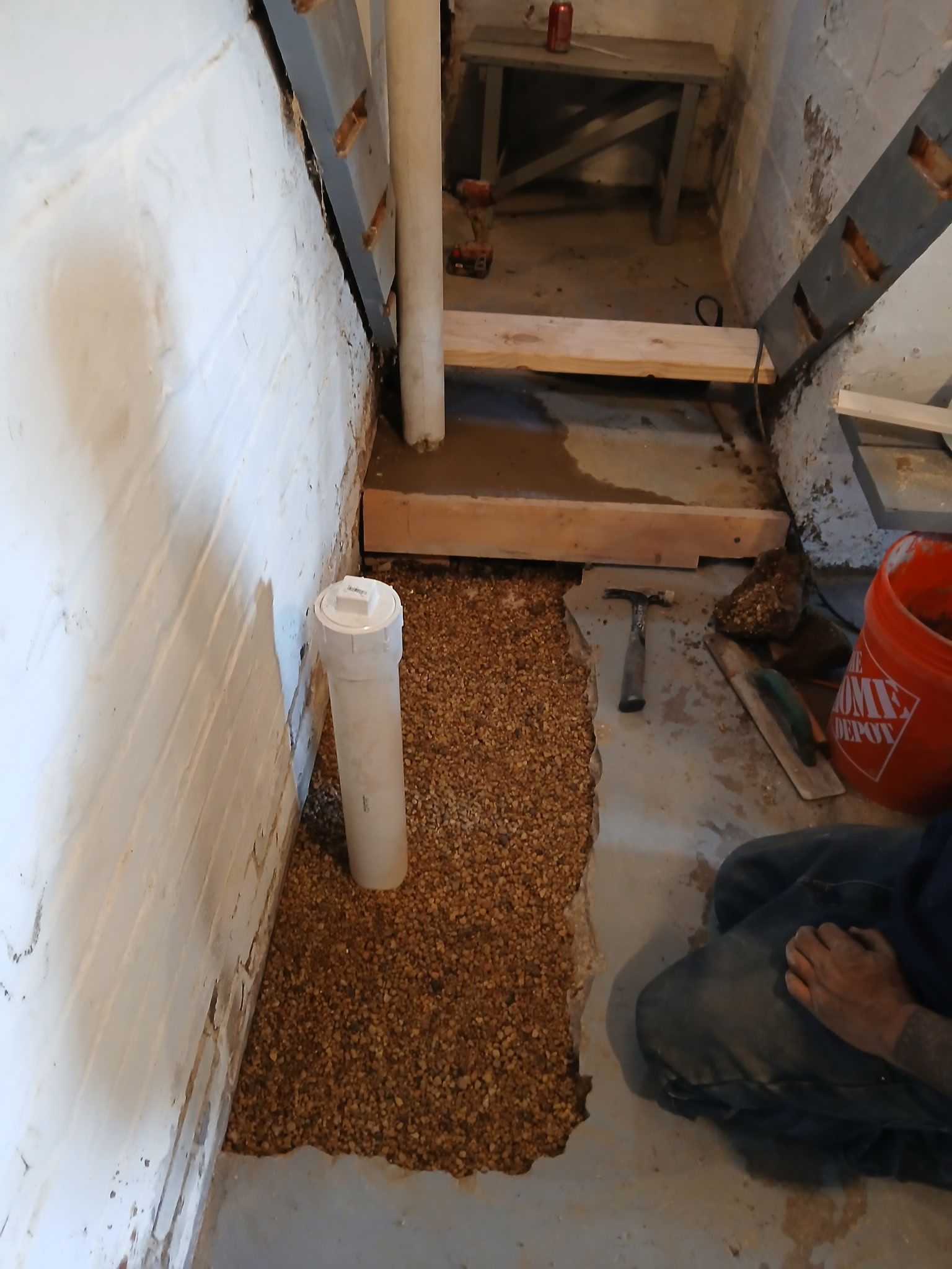 Gravel-filled trench beside a white wall, with exposed pipes and construction materials in a narrow work area