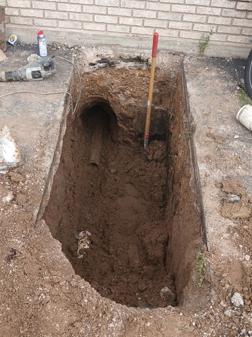 Deep rectangular excavation in a dirt yard beside a brick wall, with a shovel standing upright inside.