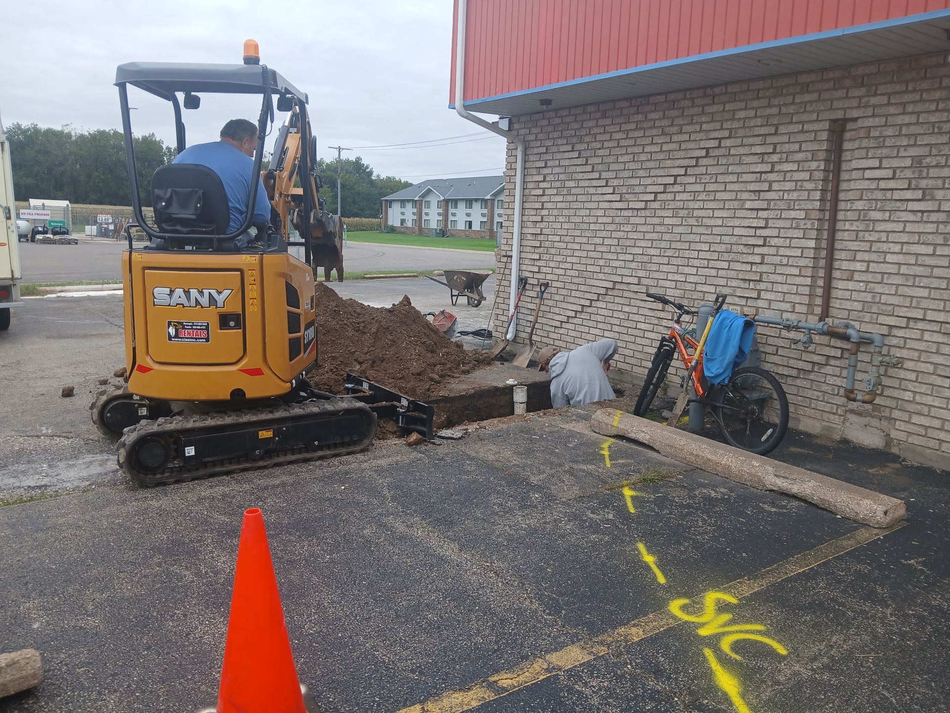 Small excavator beside a brick building, digging dirt near a parking lot cone and wheelbarrow.