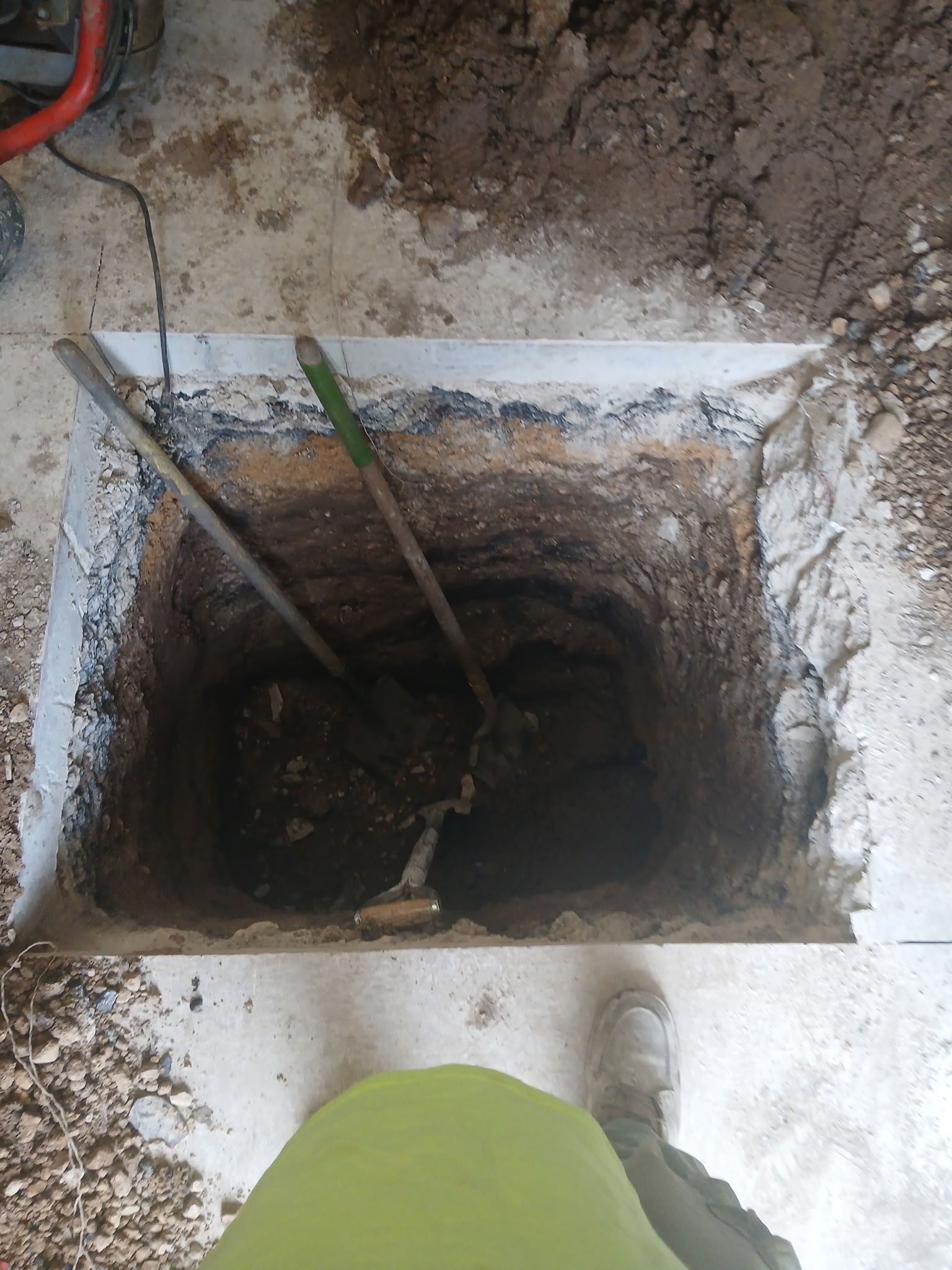 Open ground pit with dirt walls, two tools inside, and a person standing at the edge in white boots.
