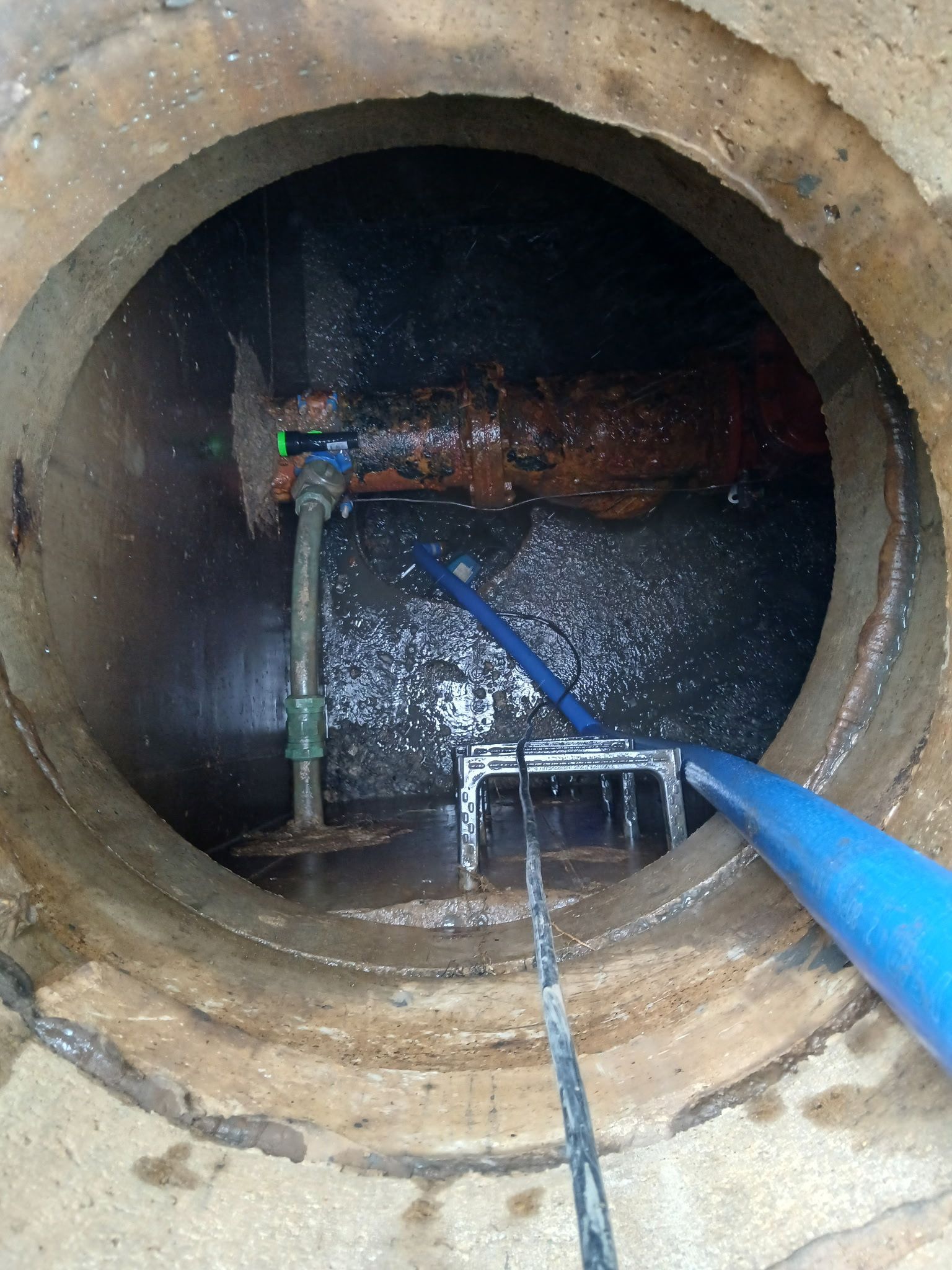 Inside a concrete manhole, pipes and cables run into dark water with a blue hose in the foreground.