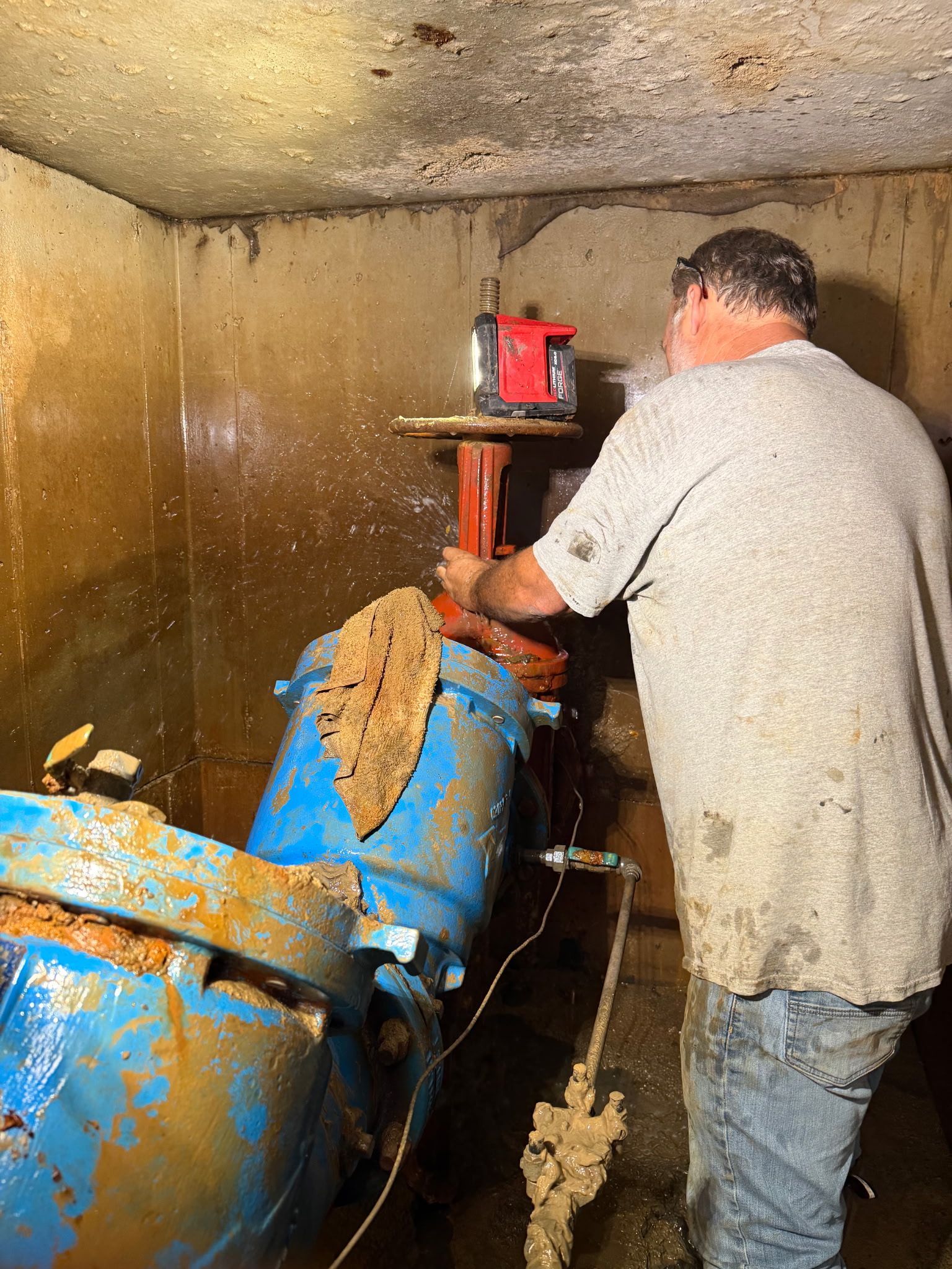 Worker beside a blue industrial machine in a cramped, dirty room, pouring material into the top funnel.