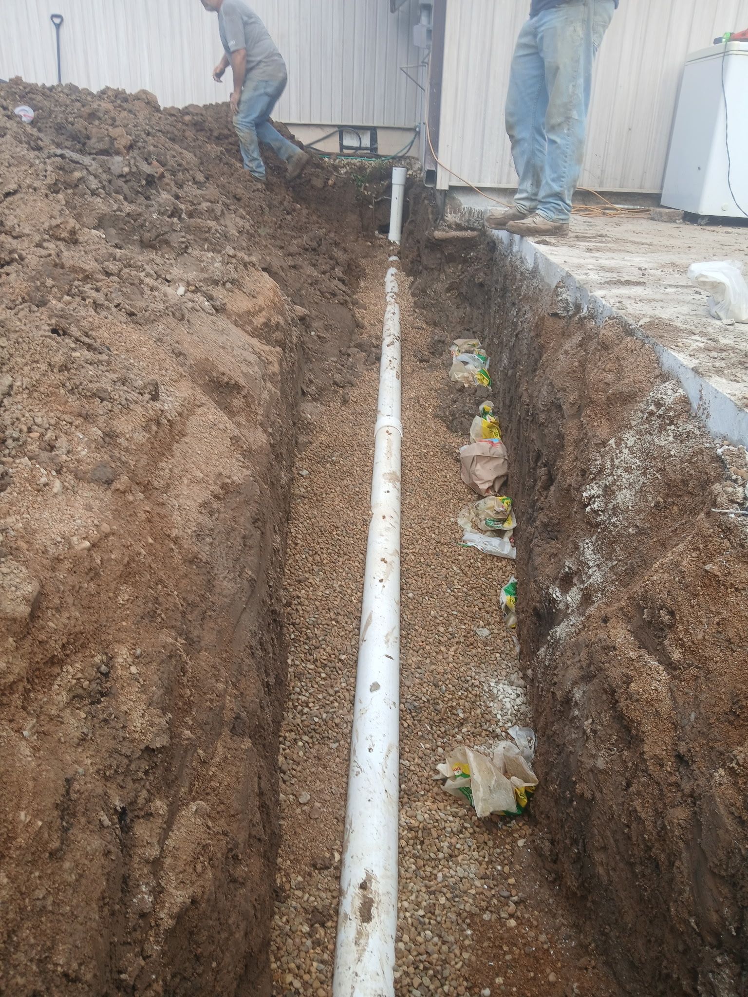 Workers installing a white pipe in a deep trench beside a building