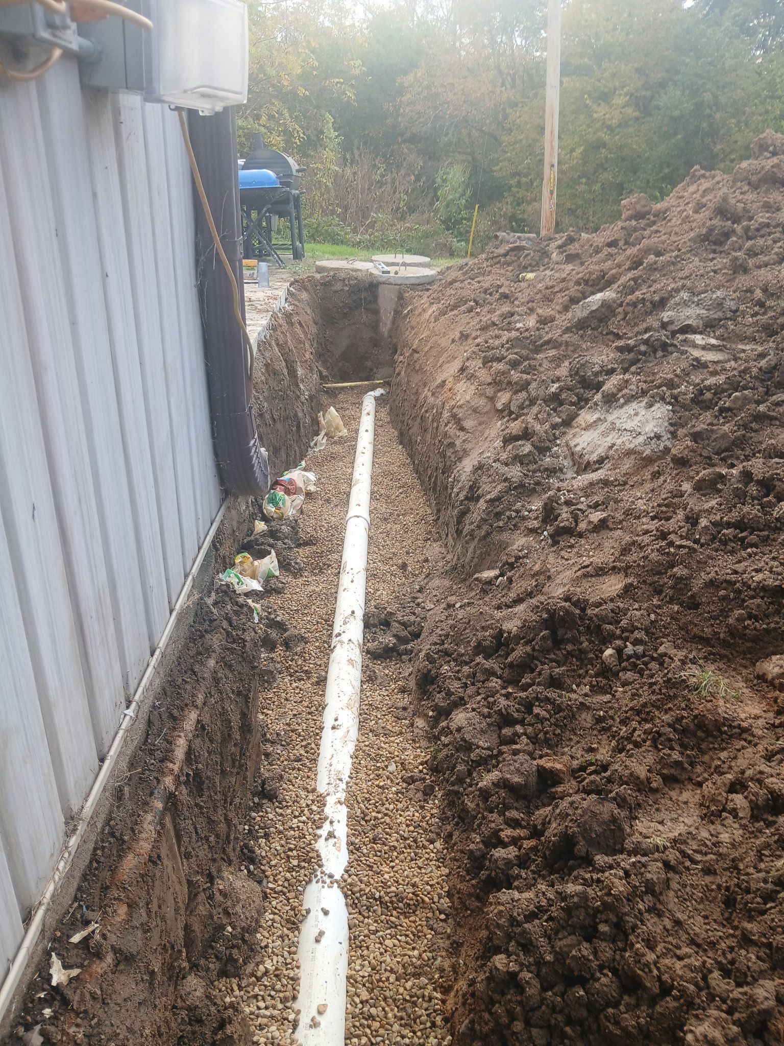 Narrow dirt trench beside a metal wall, with white pipe laid along the bottom and piles of soil on the right.