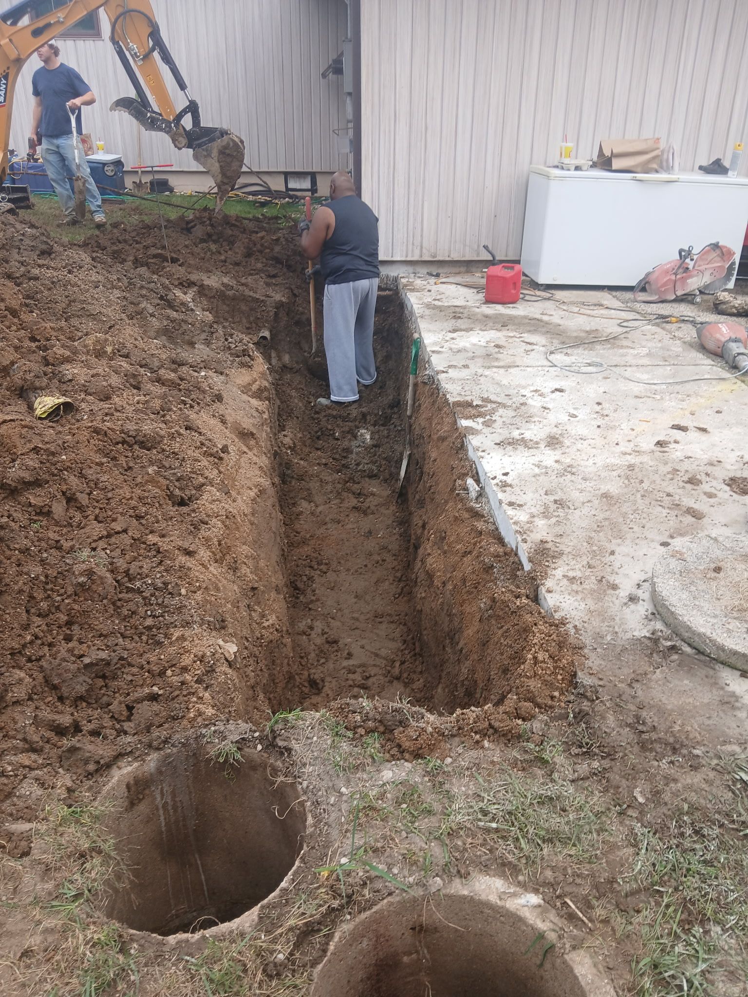 Workers digging a deep trench beside a building, with two round holes in the dirt foreground.