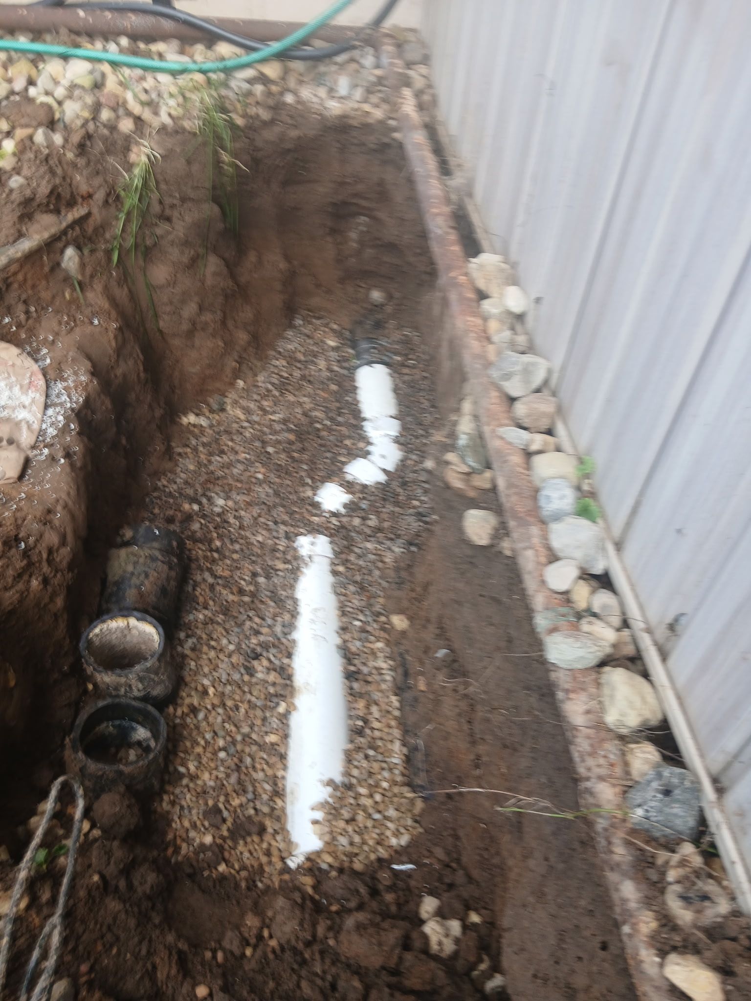 Narrow dirt trench beside a corrugated metal fence, with gravel, pipes, and white marking lines.