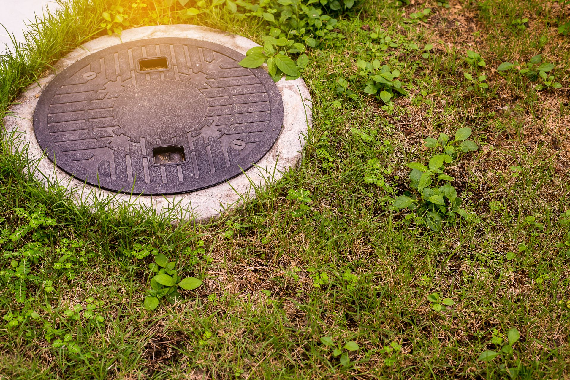 A round, dark metal manhole cover sits flush within a concrete ring surrounded by patches of grass and weeds.