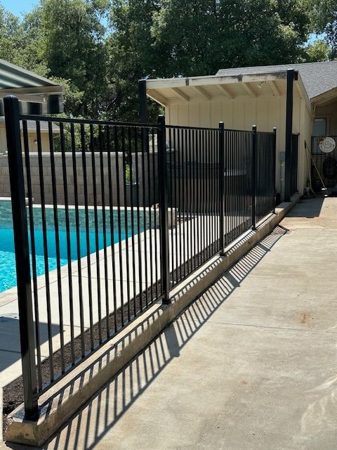 Black metal fence around a pool, on concrete, near a building with trees in background.