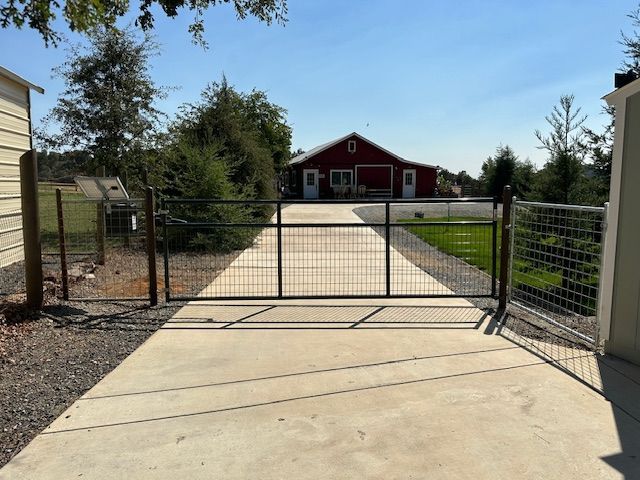 Metal gate at the end of a concrete driveway leading to a red barn under a blue sky.