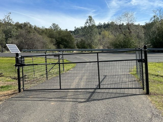 Black metal gate on a paved driveway, solar panel on left. Trees and blue sky in the background.