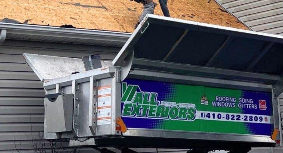 A worker on a roof using a metal chute to remove debris; a sign for All Exteriors visible.