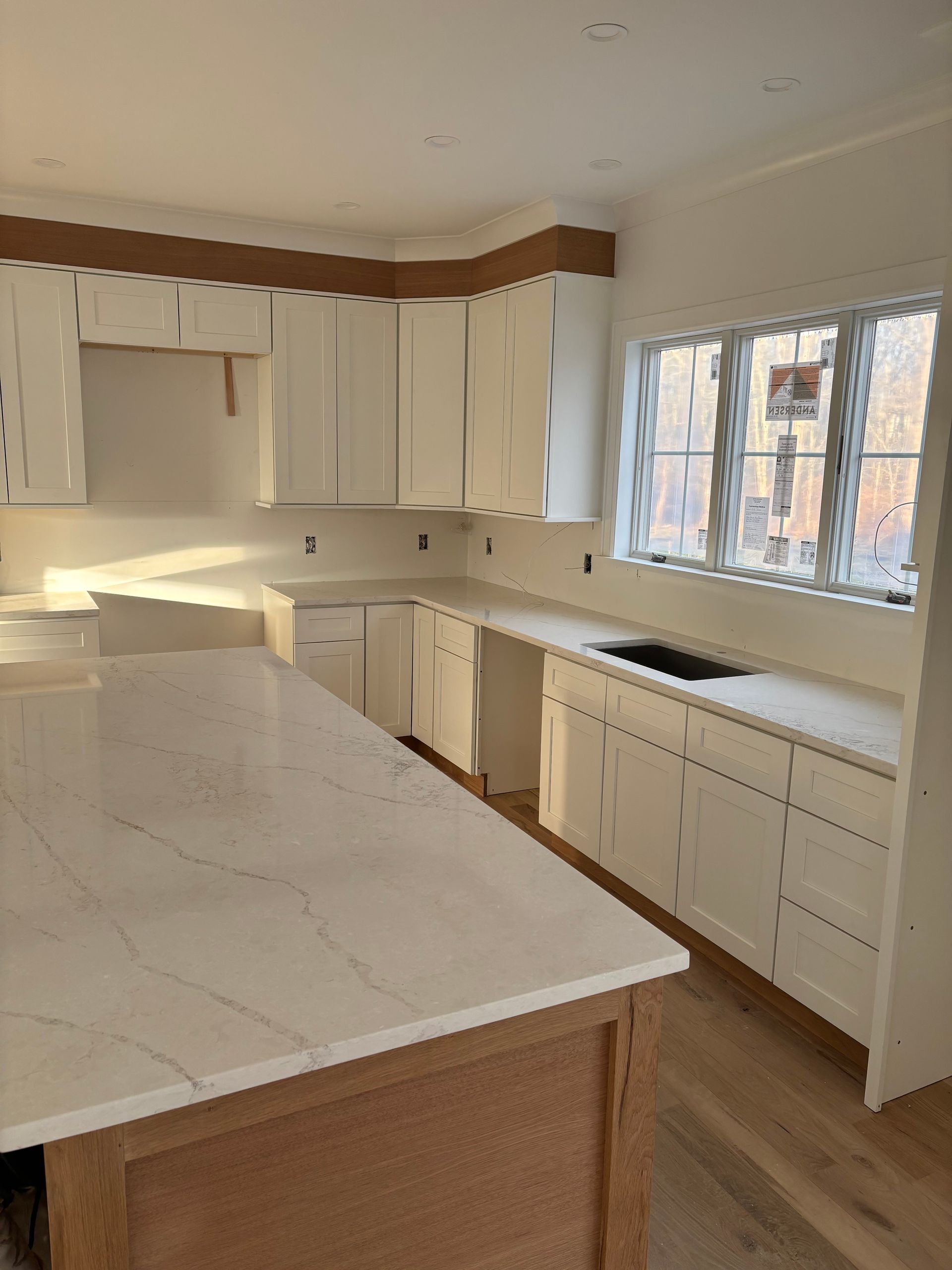White kitchen with light-colored wooden island and cabinets. Countertops have a marble-like pattern; window in the background.